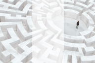 A person in formal attire stands at the centre of a large, white, three-dimensional maze. The maze walls are tall and intricate, symbolising complexity and challenges. The scene conveys a sense of isolation and problem-solving within a vast, structured environment.